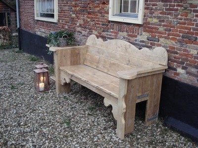 Steigerhout bank met sierlijk gezaagde planken Houten bank met een decoratieve rugleuning, geplaatst op grind bij een muur.