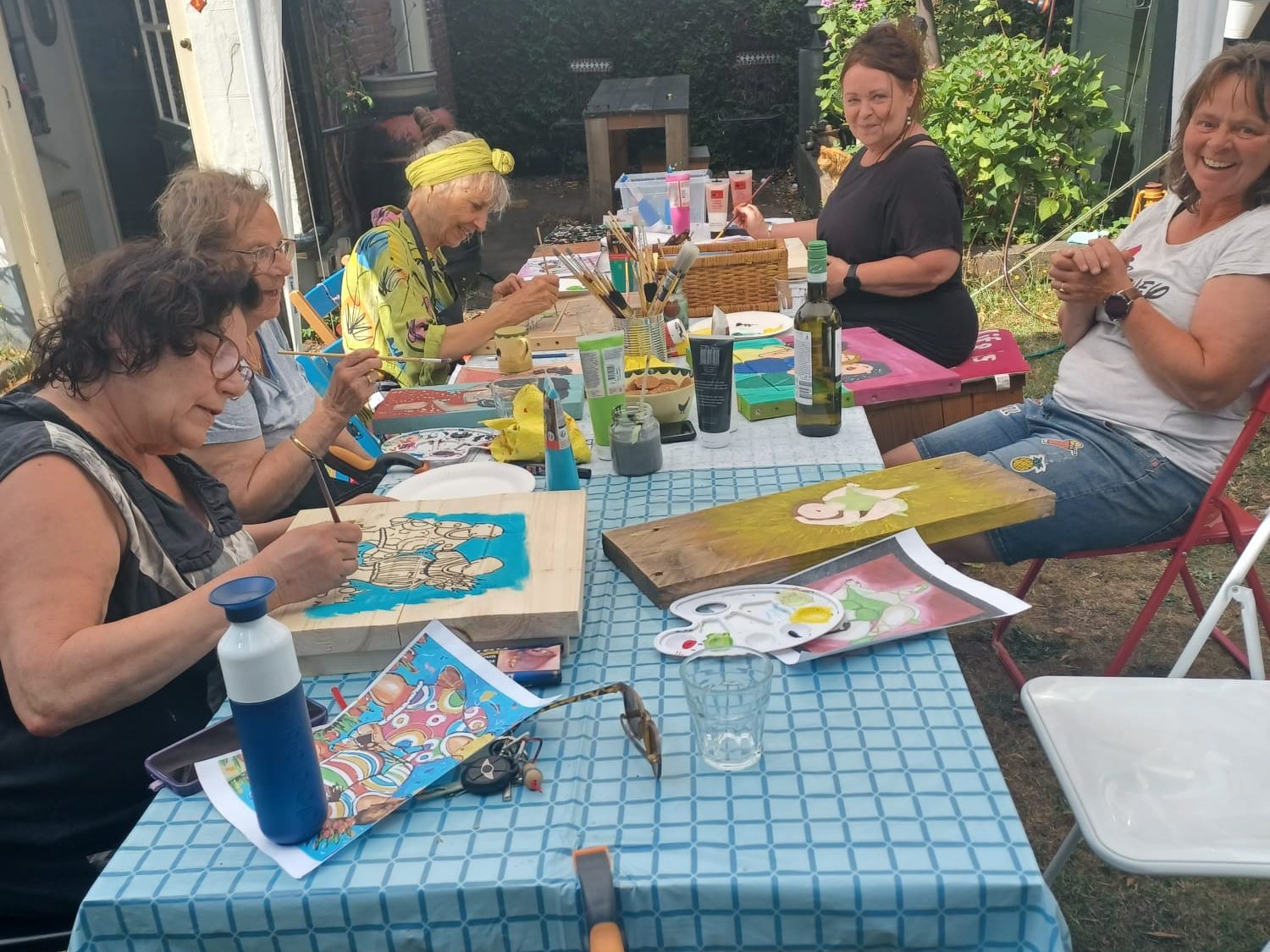 Workshop dikke dames schilderen op steigerhout Zes vrouwen aan een tafel, bezig met schilderen van dikke dames op steigerhout in een tuin.