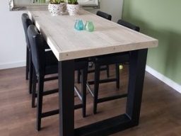 Bartafel steigerhout met stalen U poot Eettafel van hout met zwarte poten, omringd door zwarte stoelen en decoratieve planten.