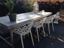 Steigerhouten tafel met halfopen blokpoot Een houten tafel met drie witte stoelen in een tuin, omringd door bloemen en planten.