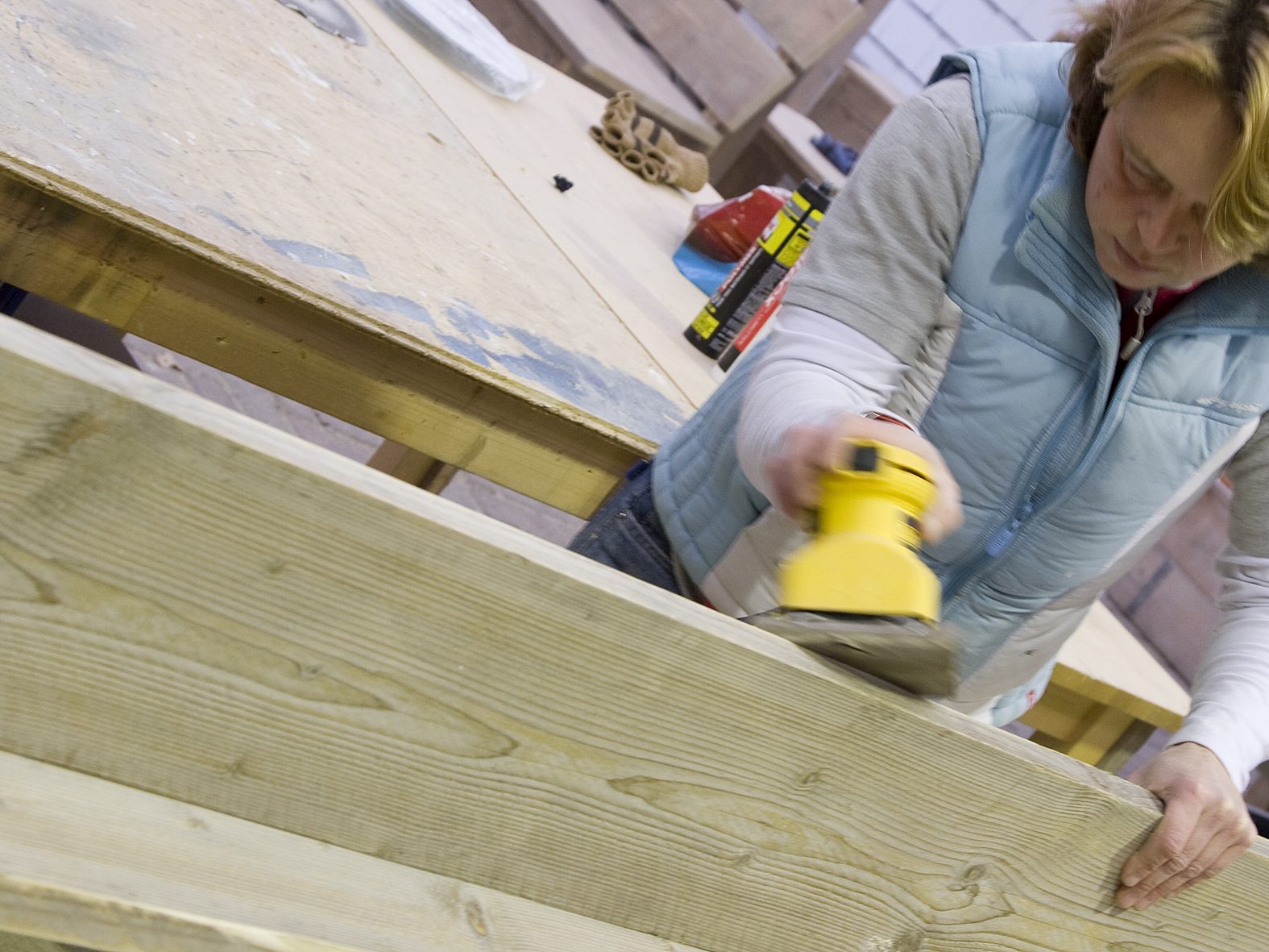Verzorging steigerhouten meubelen op locatie Een vrouw gebruikt een schuurmachine op een houten plank in een werkplaats.