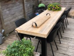 Tafel met stalen U-poot en steigerhout blad maatwerk Lang houten tafel met zes zwarte stoelen en bloempotten aan weerszijden.