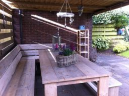 Tuintafel met hoekbank steigerhout Houten tuinset met een hoekbank onder een overkapping in een groene omgeving.